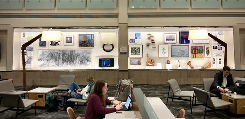 An installation image from a previous exhibition at the airport, where a display case full of visual artworks is situated behind a seating area where airport visitors are on their laptops.