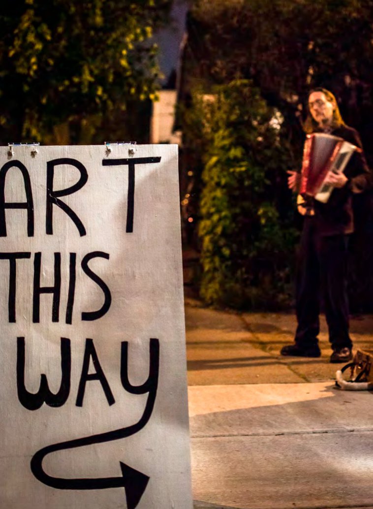 Hand-lettered Art This Way sign on a sidewalk with an accordion player in the background