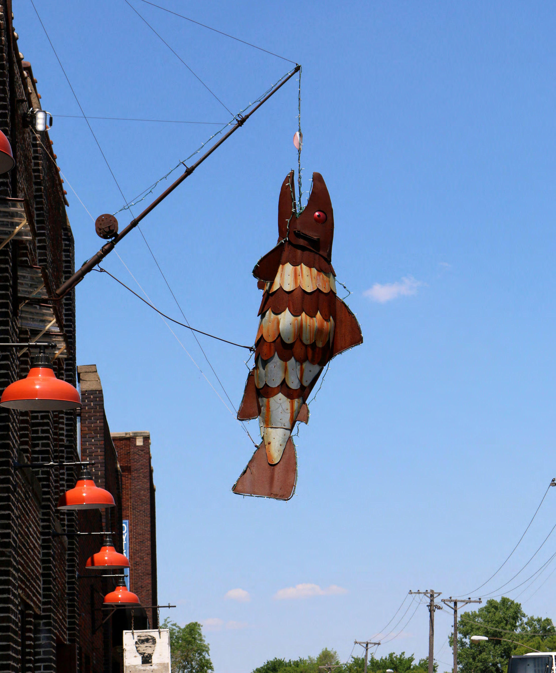 Metal fish sculpture hanging from a crane arm against a blue sky