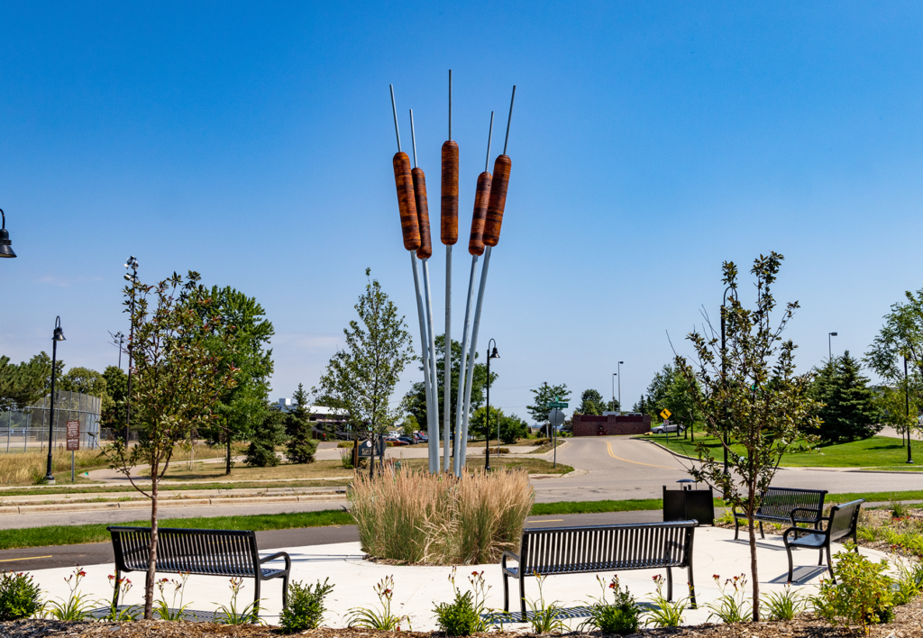 Large metal sculpture of cattails in a circular plaza surrounded by three metal benches and landscaping.