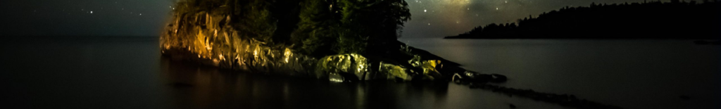 A rocky, tree-covered island illuminated at night against a starry sky over calm water.