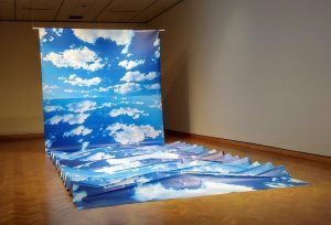 An art installation consisting of a large print of a blue sky with white clouds that hangs from the wall and flows onto the floor in accordion-like folds.