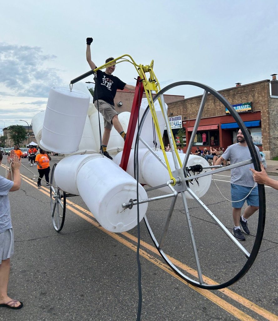 A person riding a high-wheel bicycle frame constructed from white plastic barrels in a street parade.