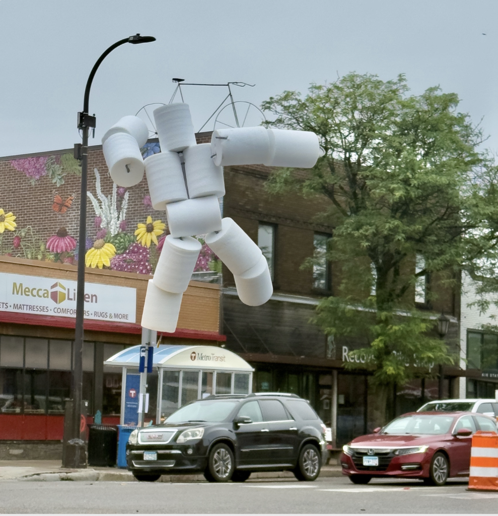 A sculpture of a human figure made of white toilet paper rolls suspended in the air above a city street.