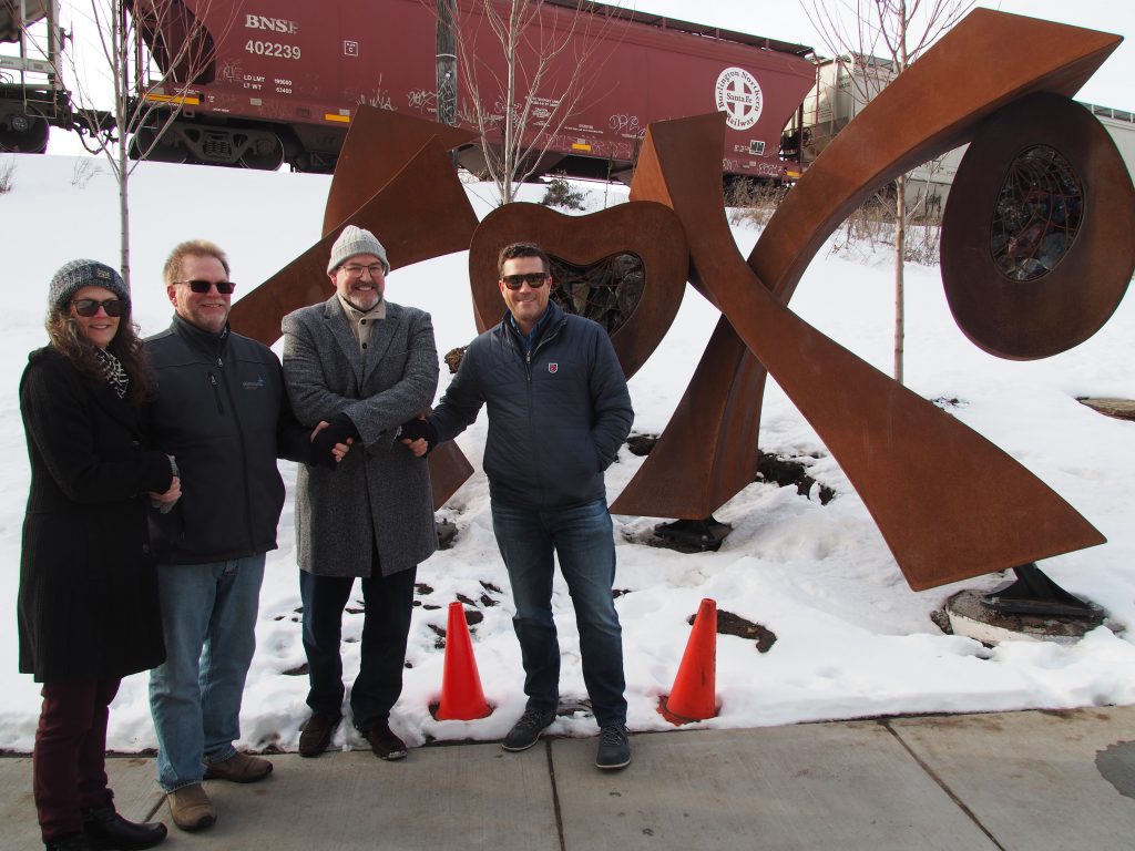 Four people pose in front of a large, rust-colored OXO sculpture in a snowy park with a passing freight train in the background.