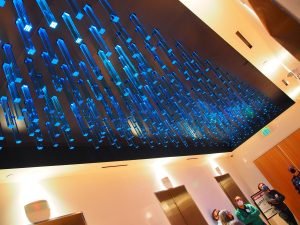 A ceiling installation of many glowing blue, rectangular light fixtures hanging over a lobby with elevators and people wearing masks.