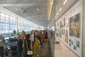 A group of people gathers in a bright hallway to view a gallery of framed artwork displayed behind a long glass partition.