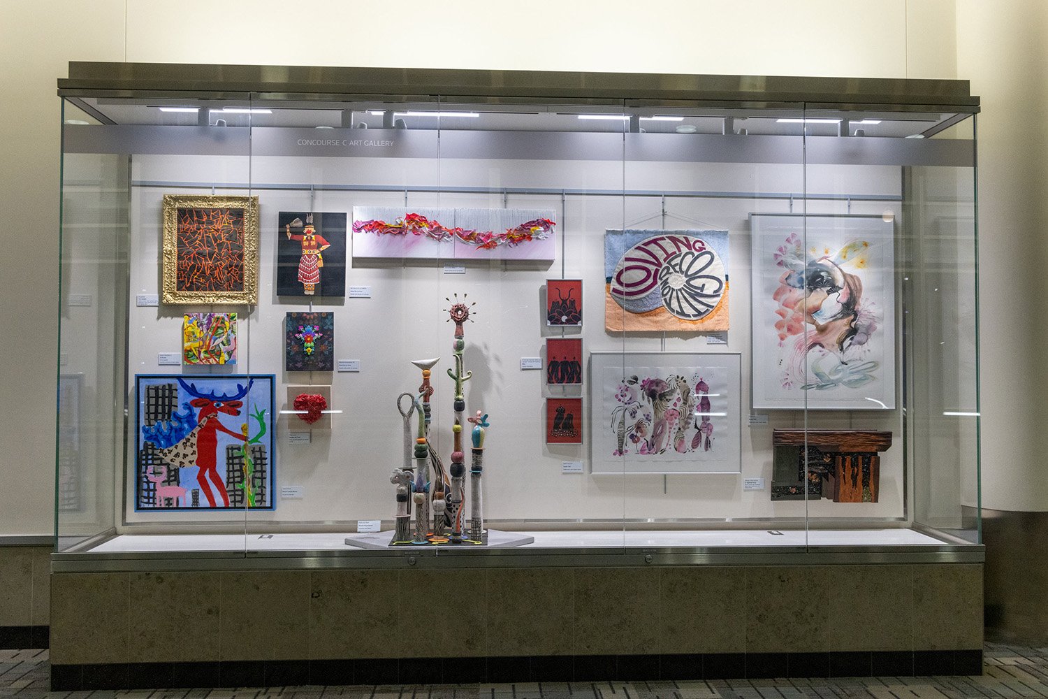 A gallery display case at an airport, featuring a collection of mixed-media artworks, including paintings, fiber art, and a central tiered sculpture.