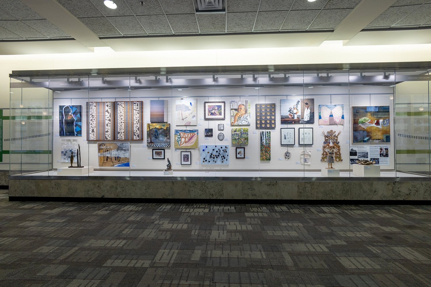 A large glass display case in a hallway containing a curated collection of various paintings, small sculptures, and framed mixed-media artworks.