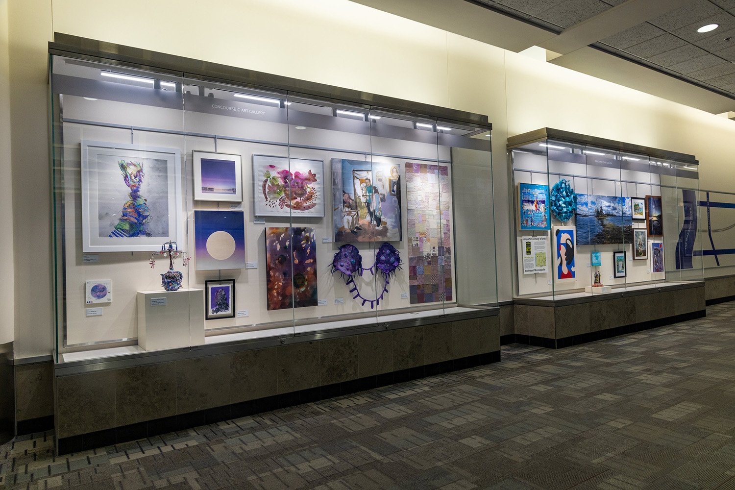 Two glass-enclosed display cases in an airport hallway showcasing various pieces of framed and sculptural artwork.
