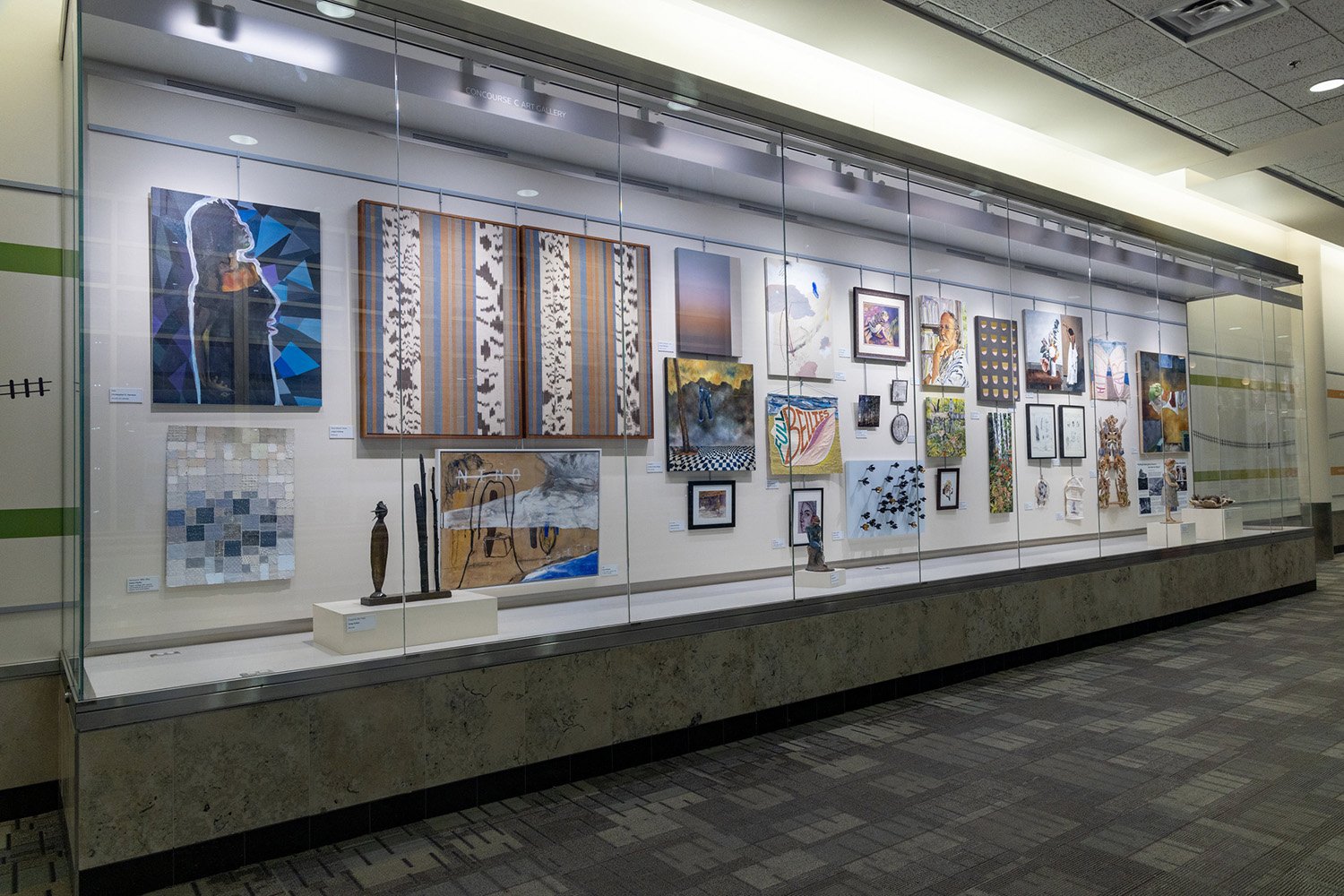A large glass display case in an airport hallway showcasing a collection of paintings, mixed-media art, and small sculptures on a tiered shelf.