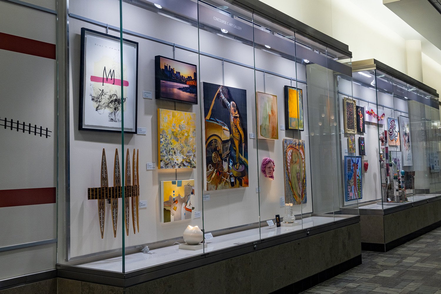 A long glass display case in an indoor hallway featuring various framed paintings, sculptures, and art pieces mounted on the wall.