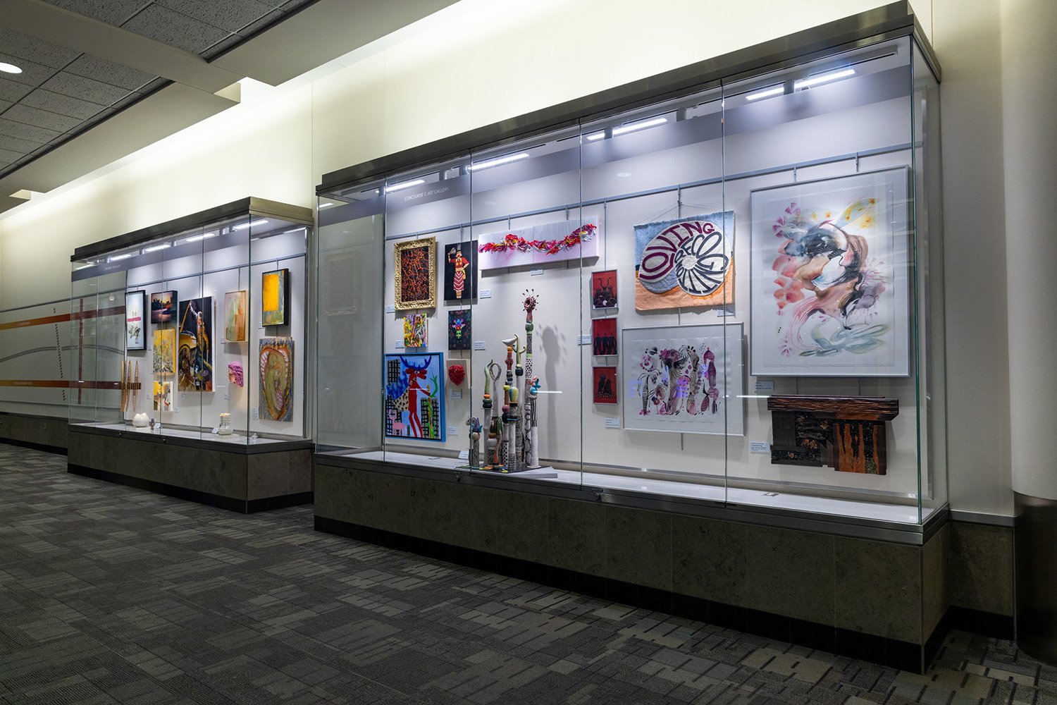 Two large glass display cases in a hallway contain a variety of paintings, mixed-media wall art, and small sculptures.