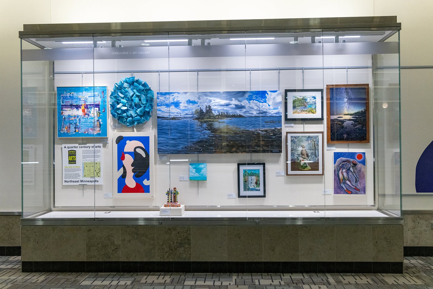 A glass display case in an airport gallery featuring various paintings, a circular blue sculpture, and a small figurine on a pedestal.
