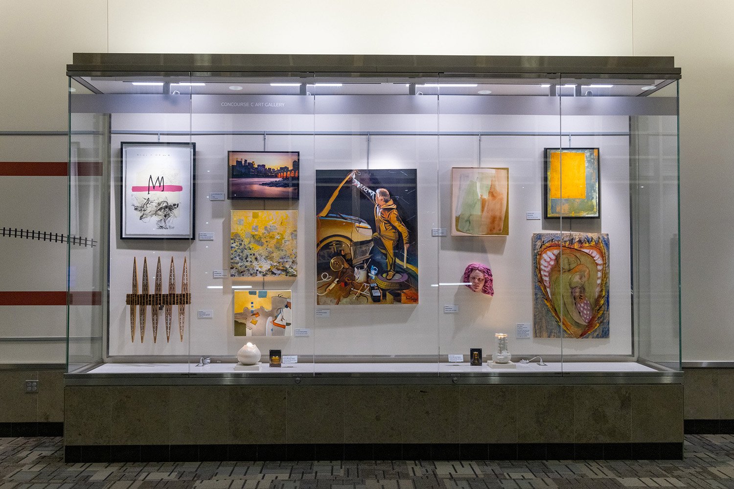 A glass display case in an airport titled Concourse C Art Gallery, containing several pieces of framed artwork and sculptures on a white wall.