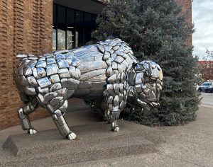 A sculpture of a bison made of reflective metal plates standing on a concrete pedestal in front of a Christmas tree.