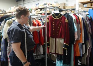 A person with a measuring tape around their neck inspects a red military-style uniform jacket hanging on a garment rack in a costume storage room.