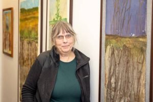 A woman with glasses and a dark zip-up jacket standing in an art gallery between paintings of landscapes.