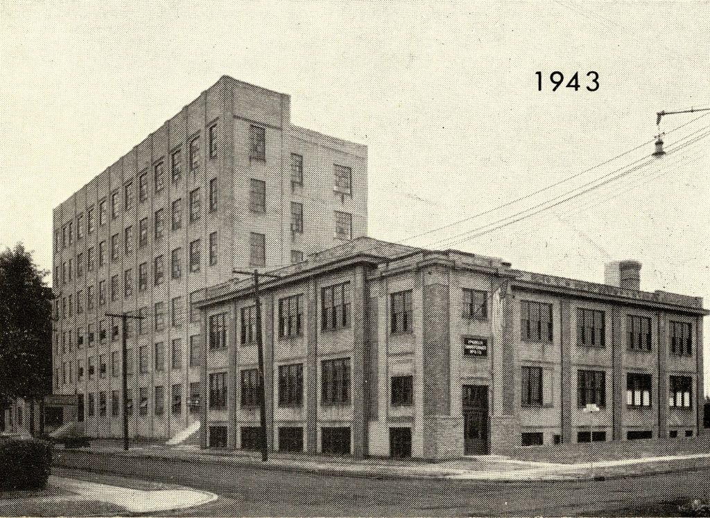 A vintage photograph from 1943 showing a multi-story industrial building attached to a shorter office building on a street corner.