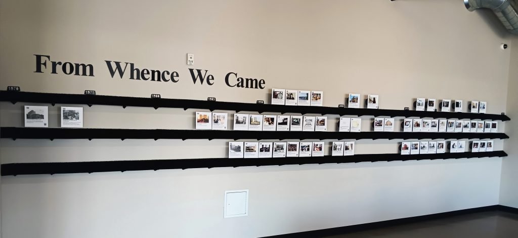 A wall display titled From Whence We Came featuring a horizontal timeline with black shelves holding multiple rows of small, framed historical photos and documents.