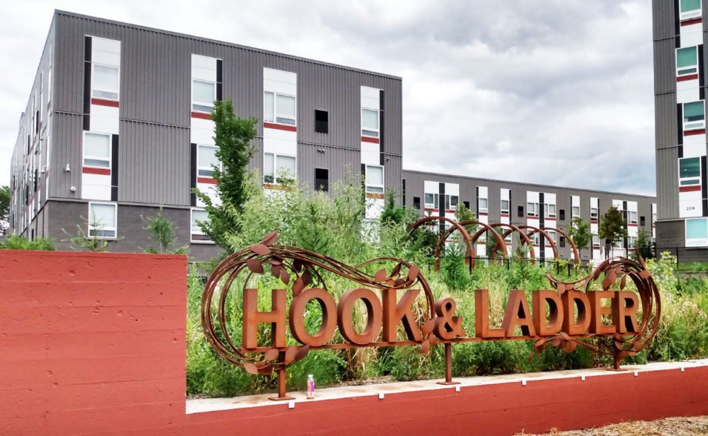 A rusted metal sign that reads Hook & Ladder in front of a modern apartment building.