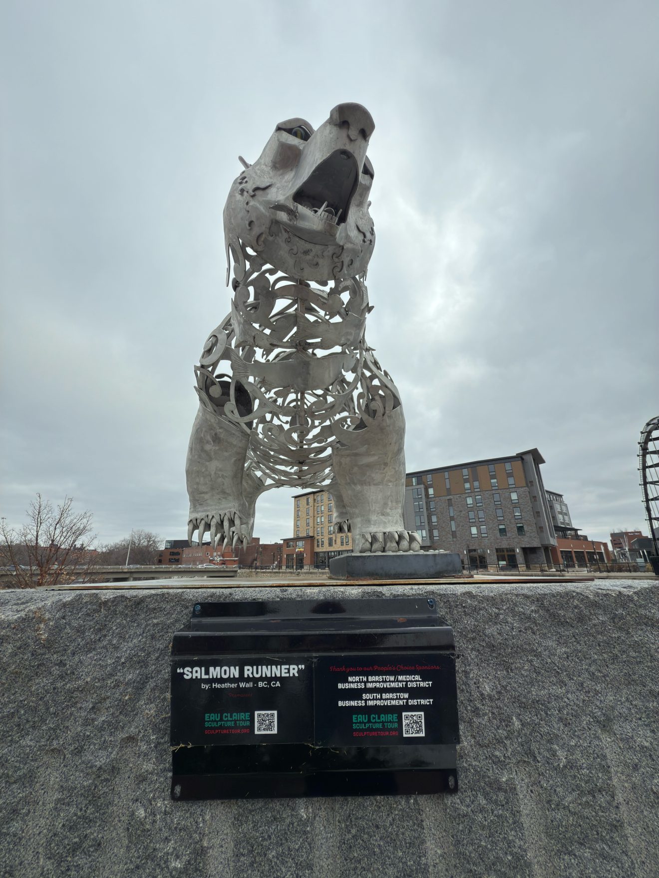 A large, silver metal sculpture of a bear titled Salmon Runner by Heather Wall stands on a granite pedestal, with a plaque displaying the artists name and QR codes below it.