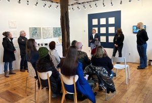 A woman speaks to a group of people seated in a bright art gallery, with square textured artworks displayed on the walls.