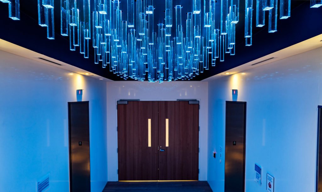 A hallway with a large, glowing blue pendant light fixture hanging from the ceiling above a pair of closed wooden double doors.