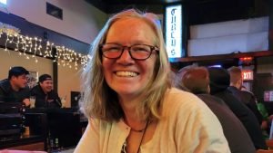 A smiling woman with shoulder-length blonde hair and glasses sits in a dimly lit bar with other patrons in the background.