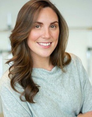 A smiling woman with long brown hair wearing a light-colored top.