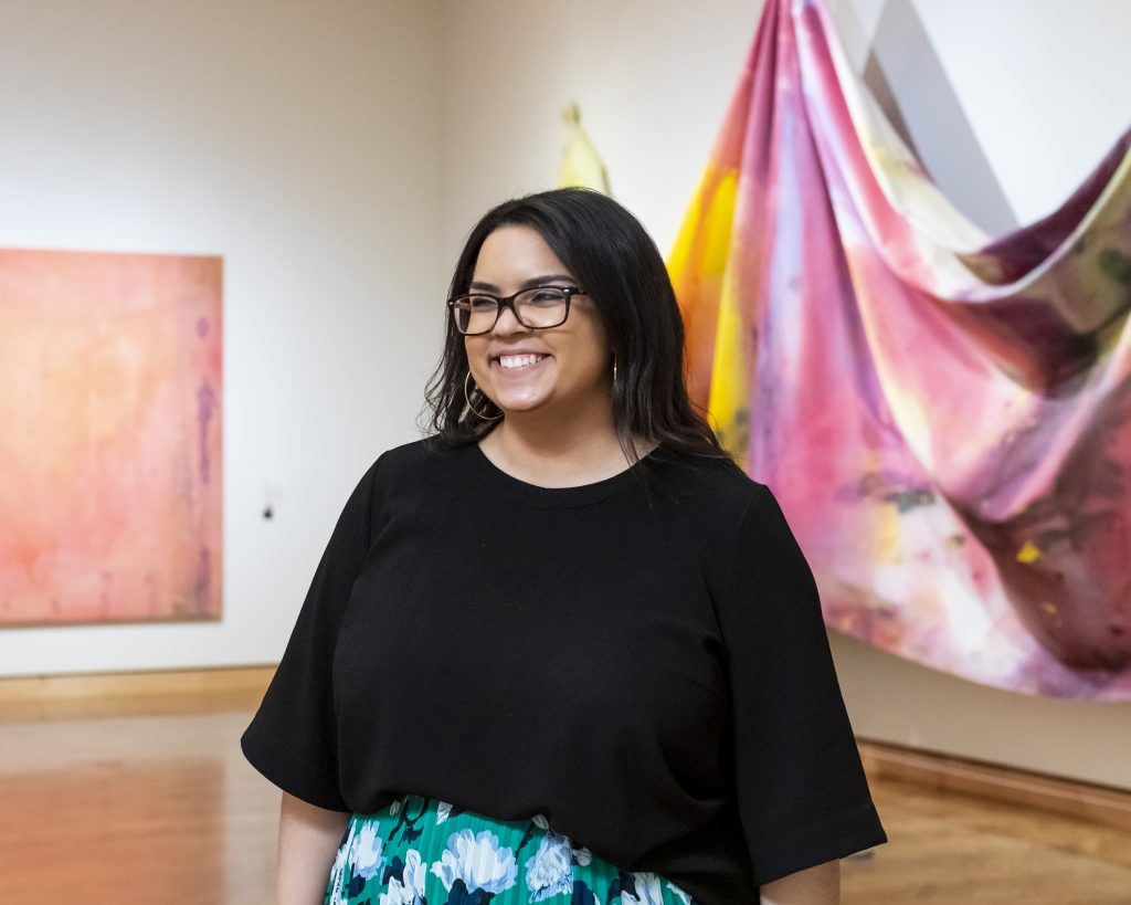 A smiling woman wearing glasses and a black top stands in an art gallery.