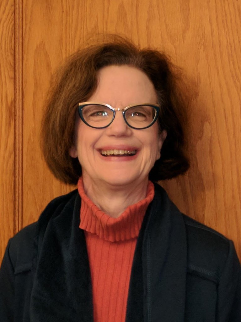 A smiling woman with brown hair wearing glasses, a red turtleneck, and a dark jacket.