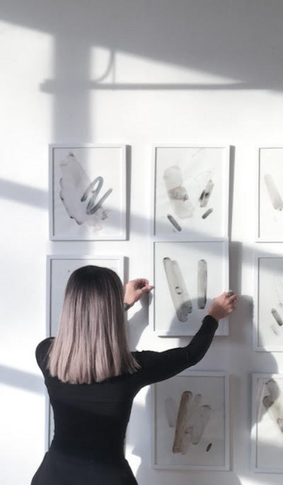 A person hanging a framed abstract painting on a white wall filled with a gallery of similar art.