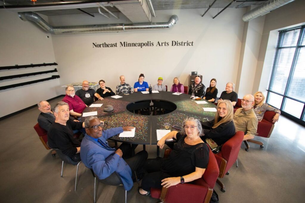 A group of people sit around a large circular conference table in a meeting room with the words Northeast Minneapolis Arts District on the wall.
