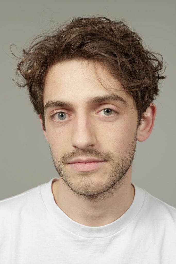 Headshot of a young man with wavy brown hair and light stubble wearing a white crew-neck shirt against a neutral background.