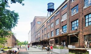 People walk and sit along a path in front of a multi-story brick industrial building with an artspace water tower on the roof.