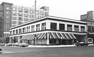 Black and white street-level view of a corner building featuring a striped awning and a tall brick building in the background.