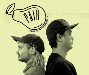 Two people wearing baseball caps stand back-to-back under the PAIR Pilot Artist in Residence logo.