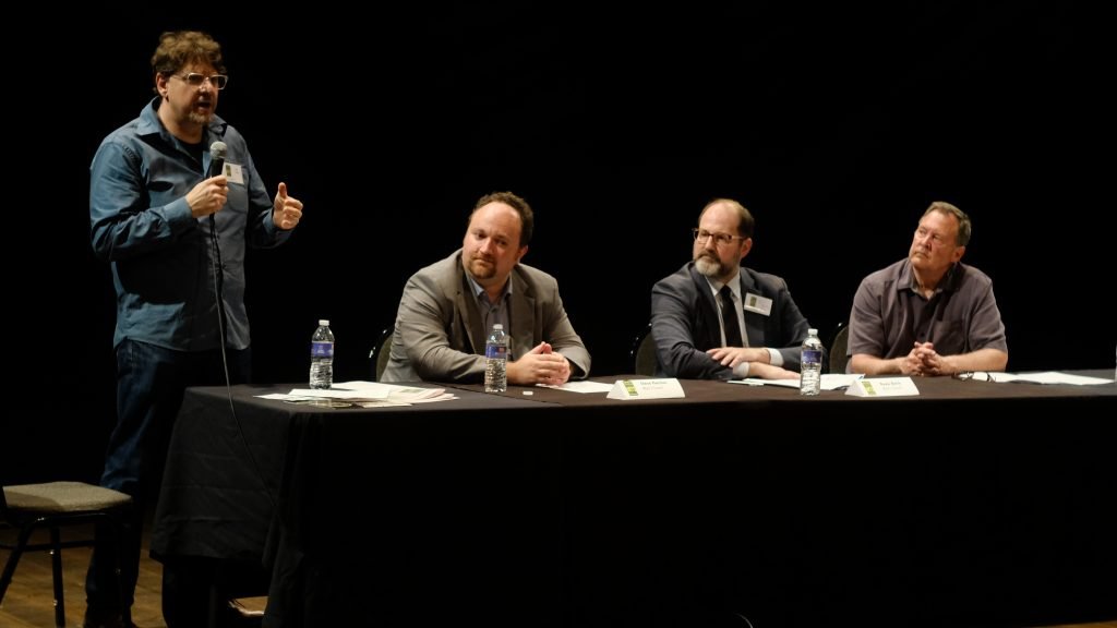 A man stands with a microphone addressing three men seated at a long table during a panel discussion.