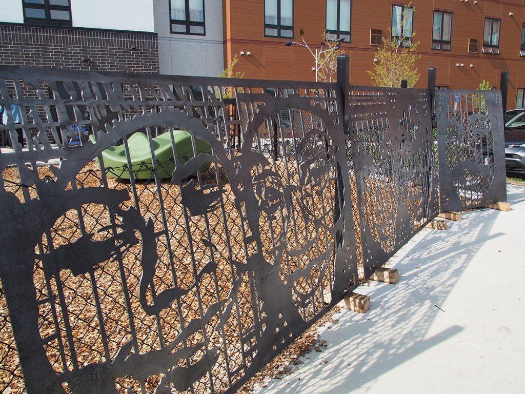 A black metal fence featuring cut-out portraits and cityscapes, installed in front of a playground area.