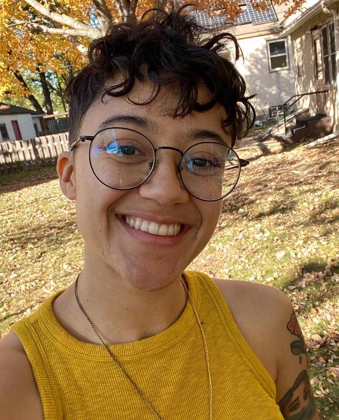 A person with short curly hair and round glasses smiles in an outdoor, leaf-covered yard.