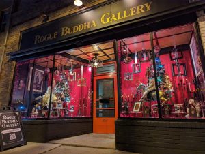 The exterior of Rogue Buddha Gallery at night, featuring a bright red storefront with two display windows filled with Christmas trees, ornaments, and art, and a sandwich board sign on the sidewalk.
