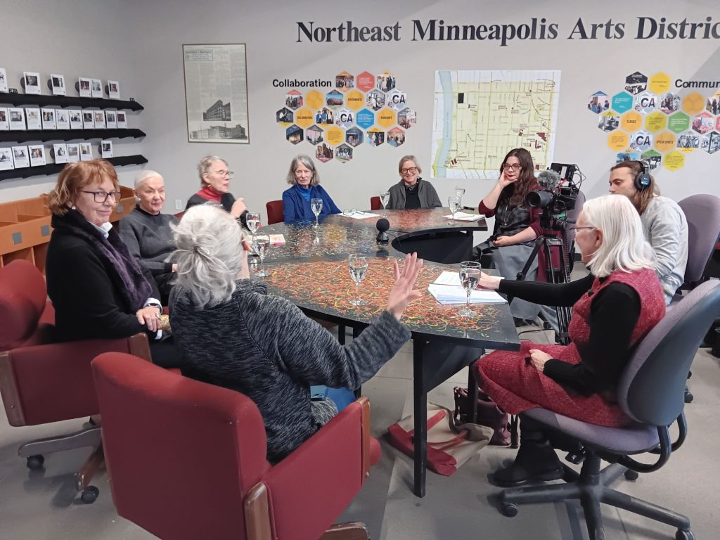 A group of eight people sit around a large, colorful conference table in a meeting room with Northeast Minneapolis Arts District on the wall, while one person records the discussion with a video camera on a tripod.