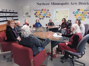 A group of eight people sitting around a large, multi-colored table in an office, with a video camera on a tripod recording the meeting.