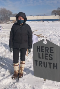 A person wearing a winter jacket, face mask, and tall boots stands in a snowy field next to a sign that reads, HERE LIES TRUTH.