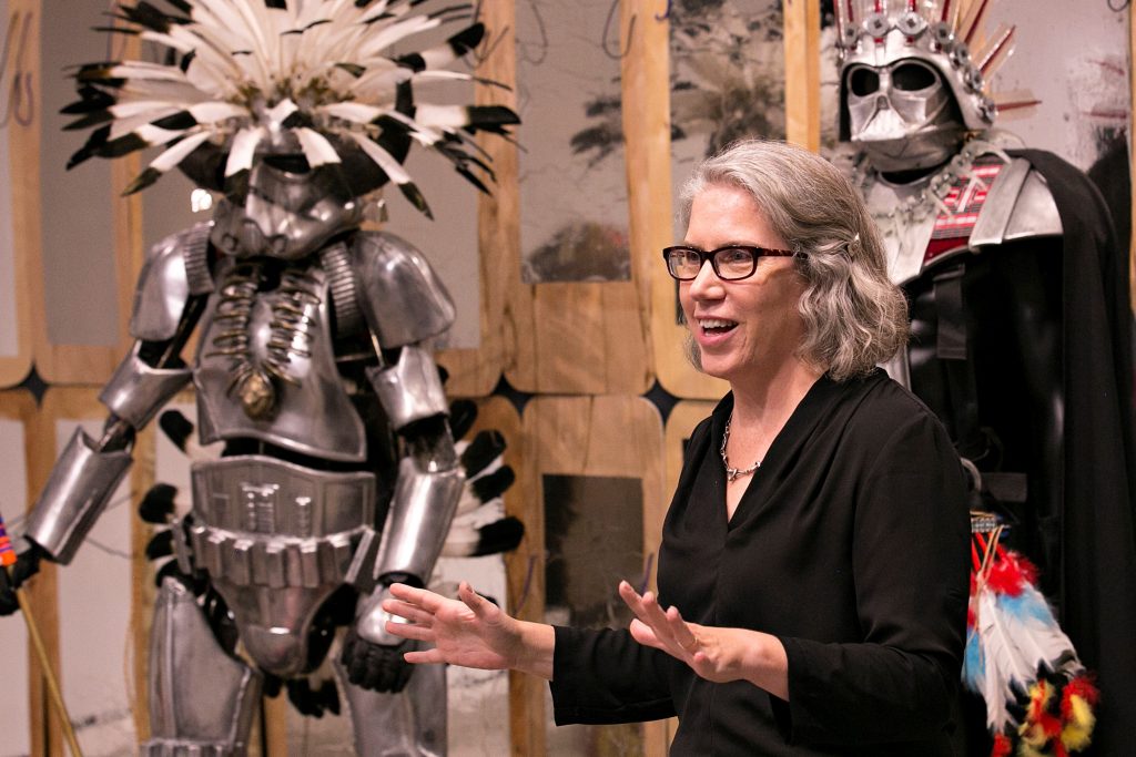 A woman with gray hair and glasses gestures while speaking in front of two figures dressed in elaborate, stylized Star Wars-themed costumes.