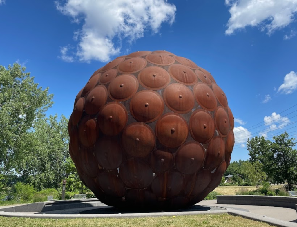 A large, spherical sculpture made of rusted metal plates with circular protrusions sits in a grassy park under a blue sky.