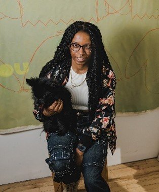 A person with long dark braids and glasses holds a small black dog while sitting on a stool.