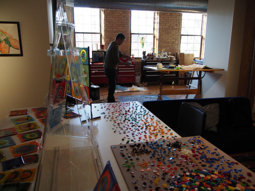 A person works at a workbench in a studio filled with colorful glass pieces, including a display rack of tiles and tables covered in assorted glass beads and gems.