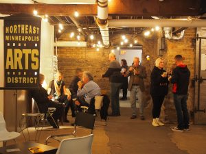 People gathered in an industrial-style space featuring a sign for the Northeast Minneapolis Arts District, illuminated by string lights.
